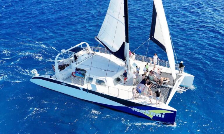 Aerial view of the catamaran "Makani," available for rent in Waikiki. The vessel is sailing on vibrant blue waters, with passengers enjoying the sun and spacious deck area, including a netted section at the front. The boat's sleek design and large white sails are prominently featured as it navigates the ocean