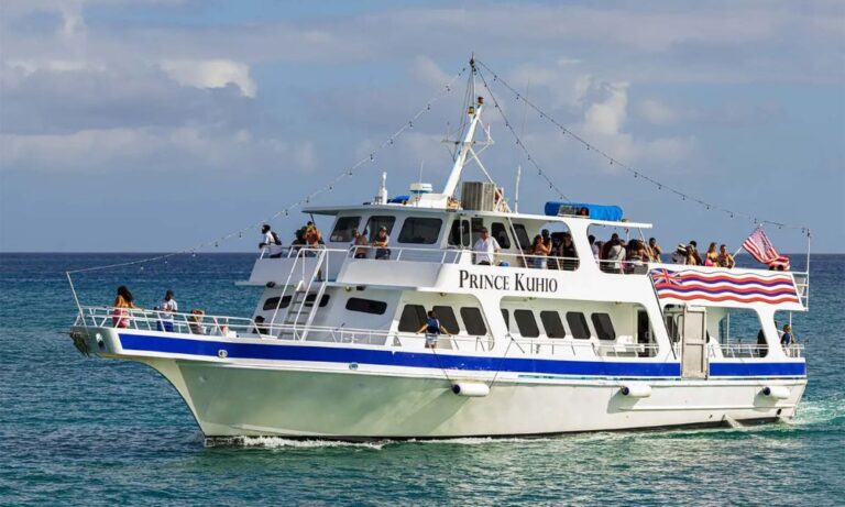 Image shows the "Prince Kuhio," a ferry available for rent in Waikiki, cruising close to the shore. The ferry is adorned with strings of lights and a Hawaiian flag, with passengers enjoying the open-air decks.