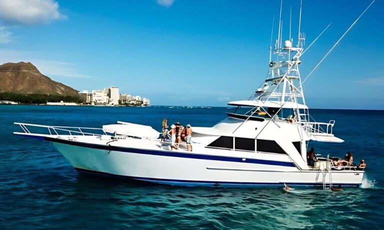 Side view of the luxury sportfisher "Striker," available for rent in Waikiki. The yacht is cruising near the shoreline, with Diamond Head and Waikiki's skyline visible in the background. Several people are on board, enjoying the sun and the spacious deck, while others swim near the boat in the clear blue ocean.