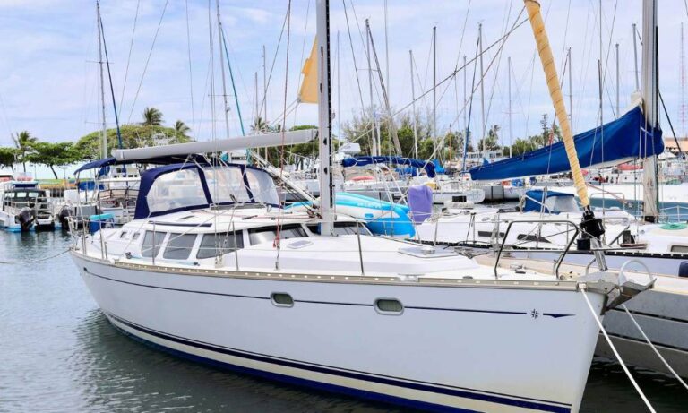 Front-side view of the charter sailing yacht "My Sunshine" docked at a marina near Waikiki. The yacht features a sleek white hull, a spacious deck with a covered cockpit, and is surrounded by other boats in the calm harbor. The backdrop includes palm trees and a partly cloudy sky, adding to the tranquil setting.