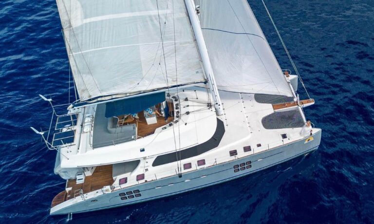 Aerial view of the catamaran "Mulan" sailing near Waikiki on deep blue ocean waters. The boat features large, white sails, a spacious deck with wooden accents, and a shaded seating area. Its sleek design is highlighted as it glides smoothly through the water.