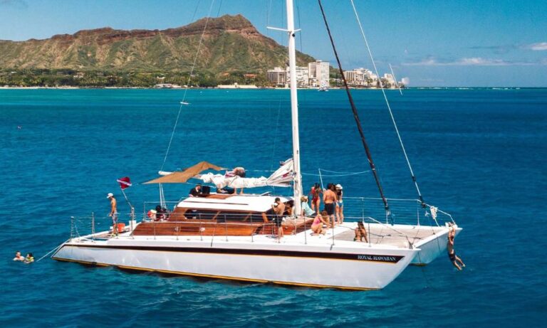 A group of people enjoying a sunny day on a large catamaran named "Royal Hawaiian" in the clear blue waters off Waikiki Beach, with the iconic Diamond Head mountain and the Waikiki skyline visible in the background. The boat is anchored, and some people are swimming in the ocean nearby.