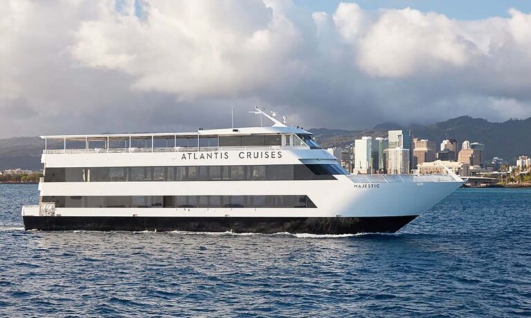 Image features the "Majestic," a large luxury yacht also available for rent in Waikiki. The yacht is seen cruising near the city skyline, with its sleek black-and-white design standing out against the ocean and clouds in the background